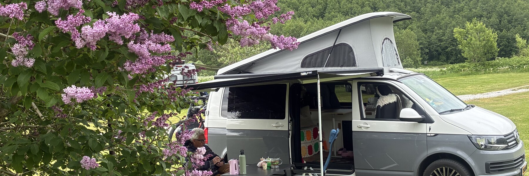 Onze VW Transporter T6 met opgeklapt dak op een Scandinavische camping, onder een bloeiende seringenboom met beboste heuvels op de achtergrond — rust en ruimte in het hoge noorden.